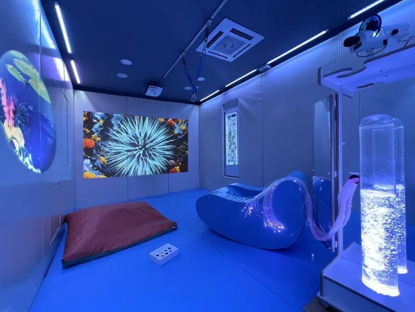A sensory room lit in blue, with a bubble tube, fibre-optic lights and soft seating