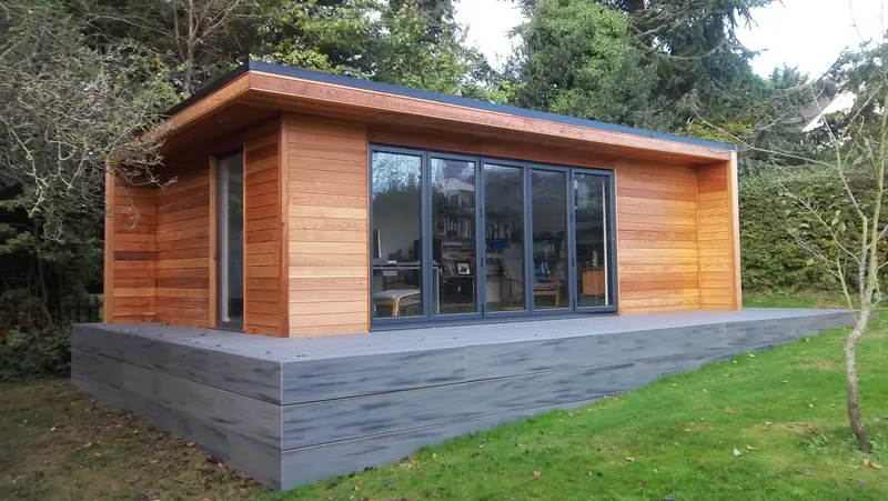 Cedar-clad garden office with grey bi-fold doors and a desk visible inside, raised on composite decking