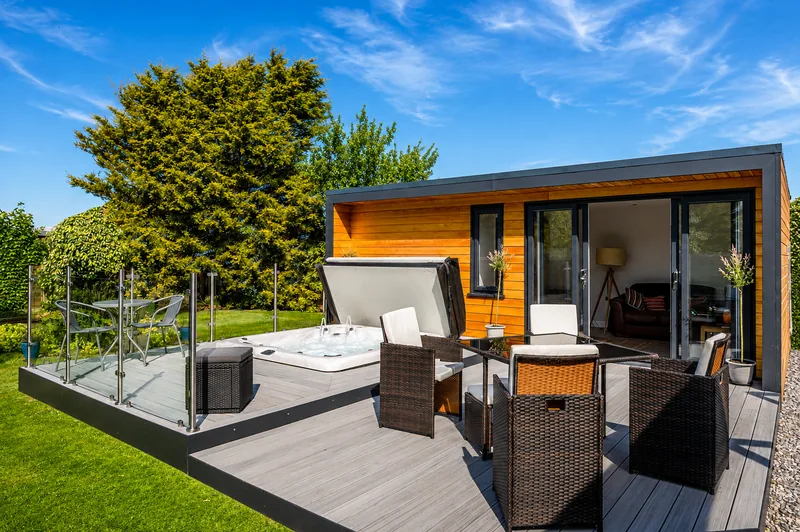 Hot tub and rattan seating on composite decking behind a glass balustrade, the garden room's sliding doors open beside them