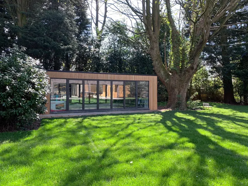 Long slatted-larch garden office with a full run of bi-fold doors, beneath a mature tree on a sunlit lawn