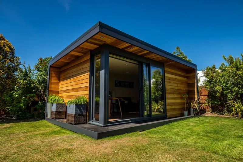 Cedar garden room with anthracite frame, recessed porch and planters on dark composite decking under a blue sky