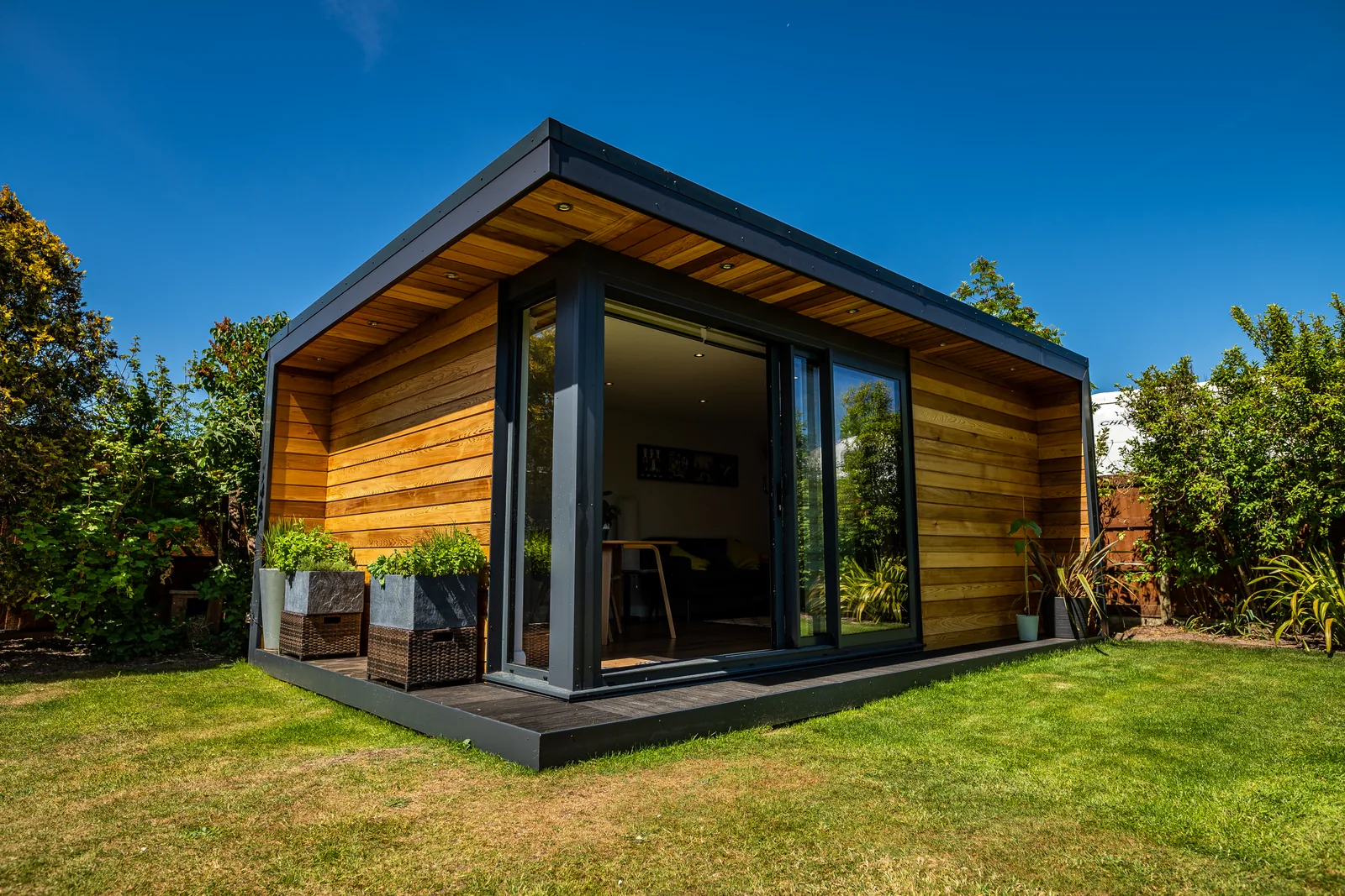 Cedar-clad garden room with an anthracite frame and a deep roof overhang, sliding doors open onto the lawn