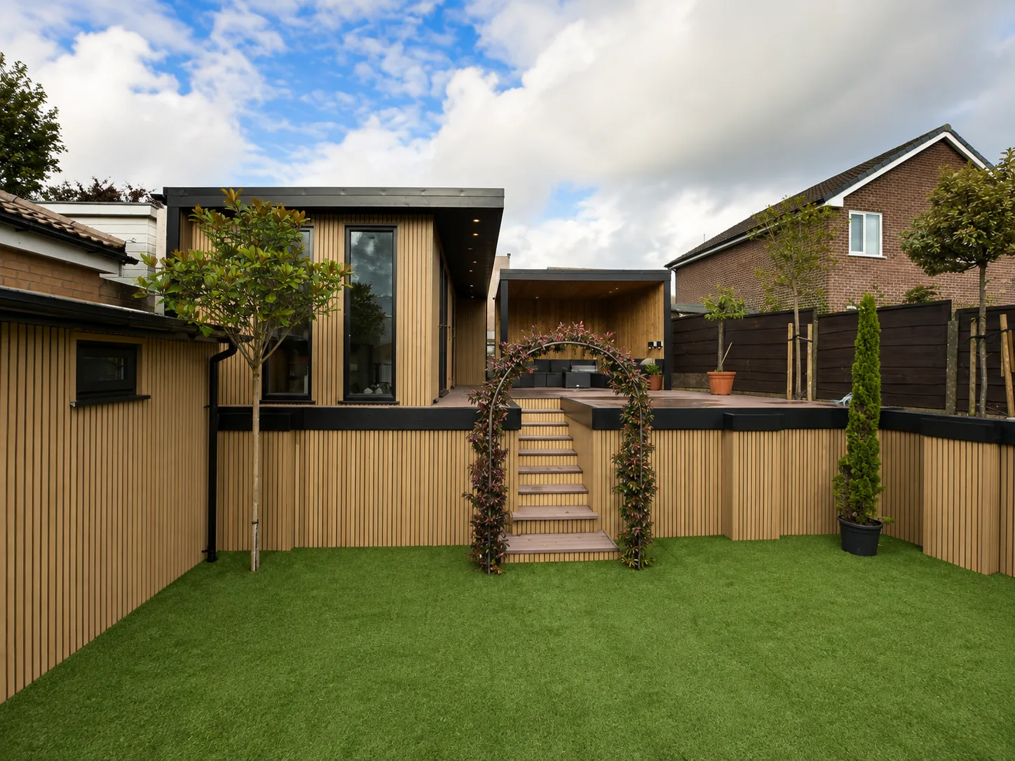 Custom build seen across an artificial lawn, a rose arch and steps leading up past the slatted timber screen