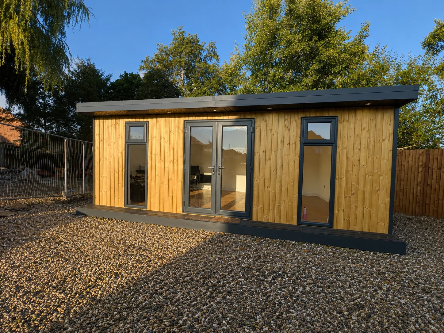 Long light-cedar garden room with a charcoal fascia, French doors and windows, on gravel beneath a willow