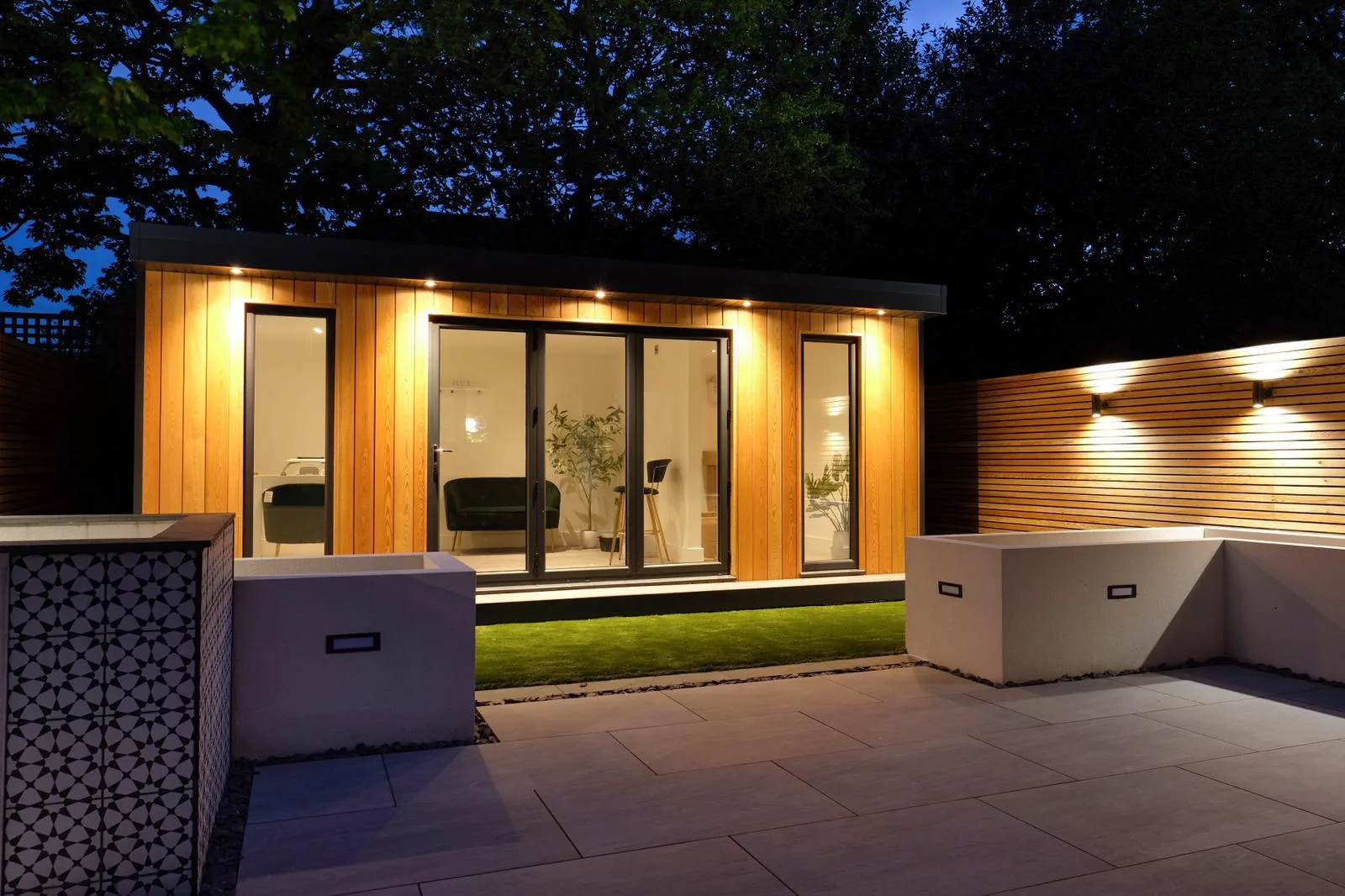 Cedar garden room at dusk, warm light spilling from the glazing across a paved terrace and rendered planters