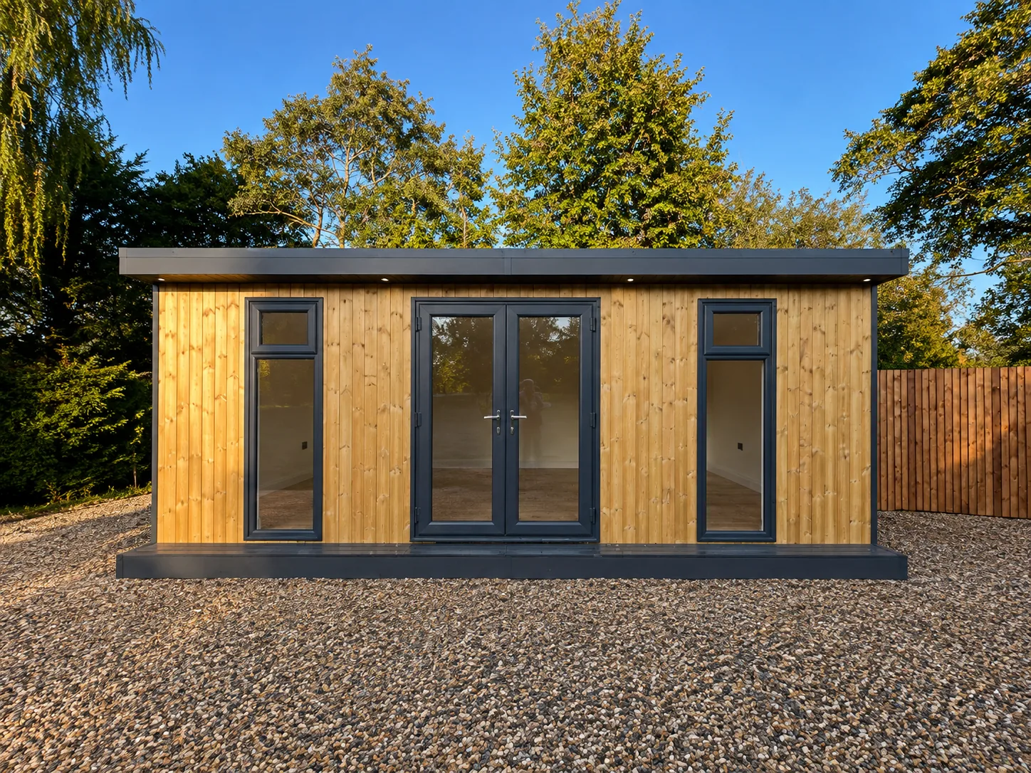 Long pale-cedar garden room with charcoal frames and French doors, on gravel with mature trees behind
