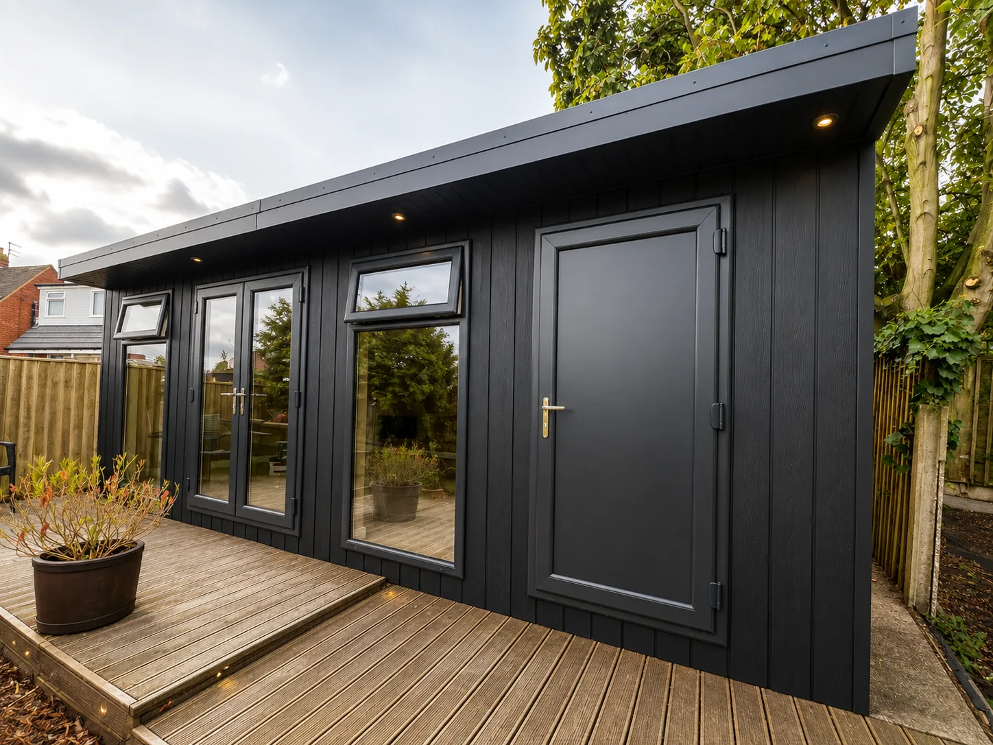Dark-clad garden room with a grey door and a run of windows, on timber decking with a potted olive